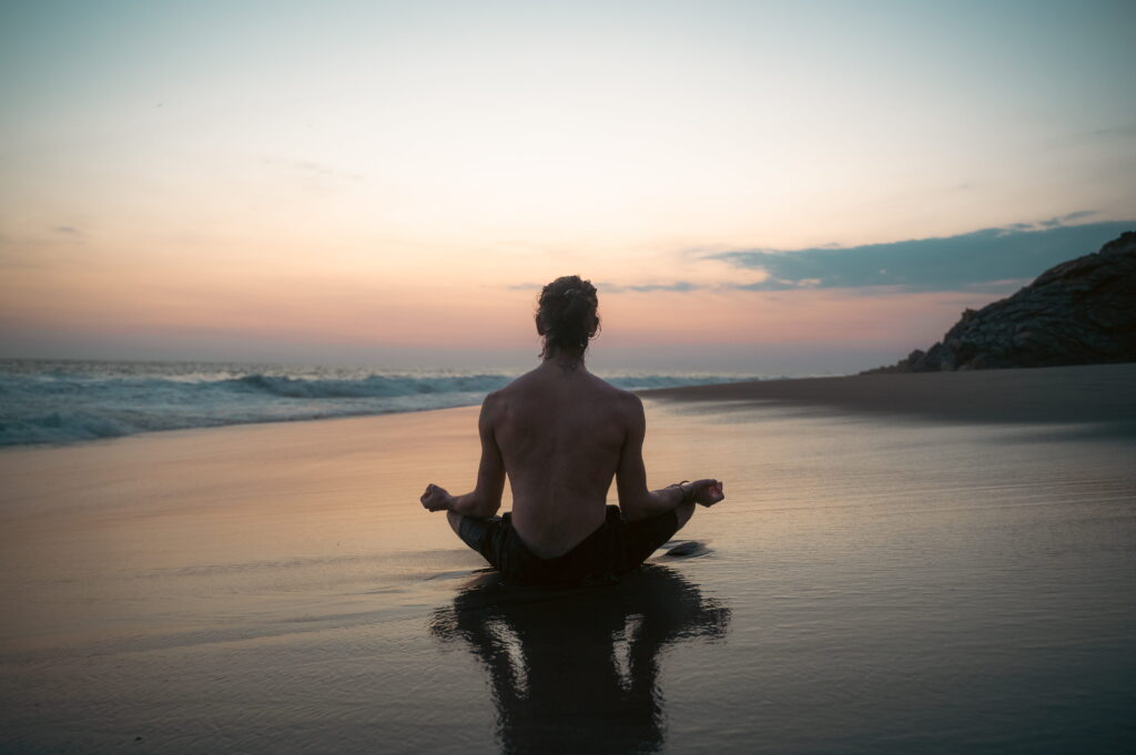 Nicolas Loewer meditating cross-legged on the beach, facing the sunset—a symbol of presence and embodiment