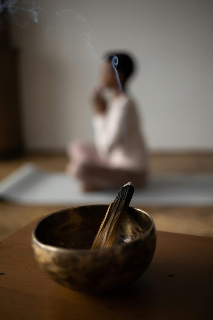 Singing bowl with Palo Santo in the foreground, with a person in a yoga pose in the background as a symbol of mindfulness, grounding, and presence